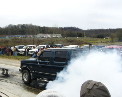 2007 Ford Mustang - A dark blue pickup truck emits a large cloud of smoke during an event, with other vehicles and spectators in the background.