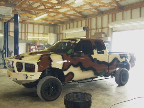 2007 Ford Mustang - A pickup truck with a camouflage paint scheme is parked inside a garage with a lift.