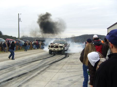 2007 Ford Mustang - A truck performs a burnout, kicking up smoke and leaving tire marks on the asphalt as onlookers watch.