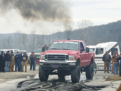 2007 Ford Mustang - A lifted red Ford F-250 truck emits a large cloud of black smoke during a burnout, with spectators watching.