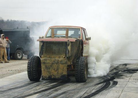 2007 Ford Mustang - Modified pickup truck with large tires blowing white smoke on a track, another truck in background.