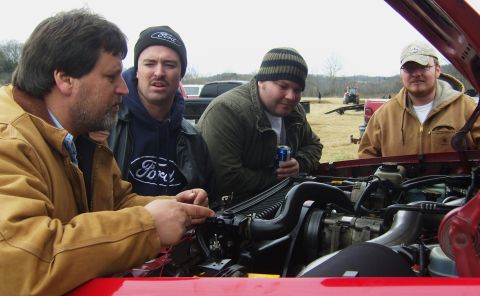 2007 Ford Mustang - Four men examine the engine bay of a red vehicle outdoors, with other vehicles and a tractor visible in the background.