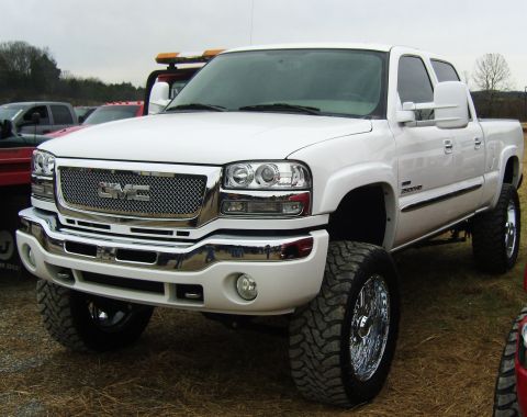 2007 Ford Mustang - A lifted white GMC Sierra pickup truck is shown from a 3q-front angle, a prominent chrome grille and large off-road tires.
