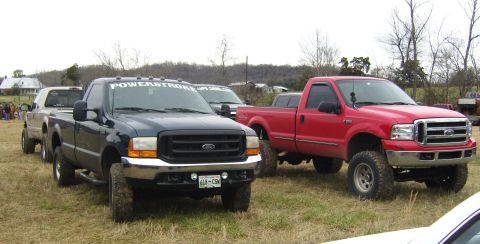 2007 Ford Mustang - Two lifted Ford trucks, one dark blue and one red, are parked on a grassy field with houses in the background.