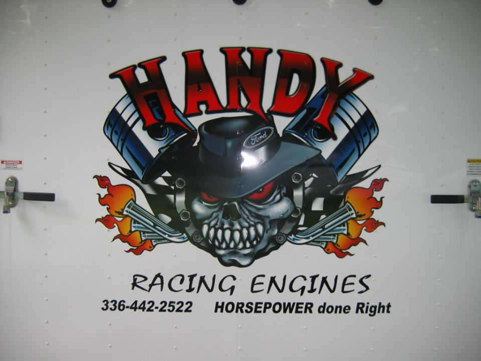 White trailer door with a graphic of a skull wearing a Ford hat, surrounded by pistons and flames, with text 'HANDY RACING ENGINES'