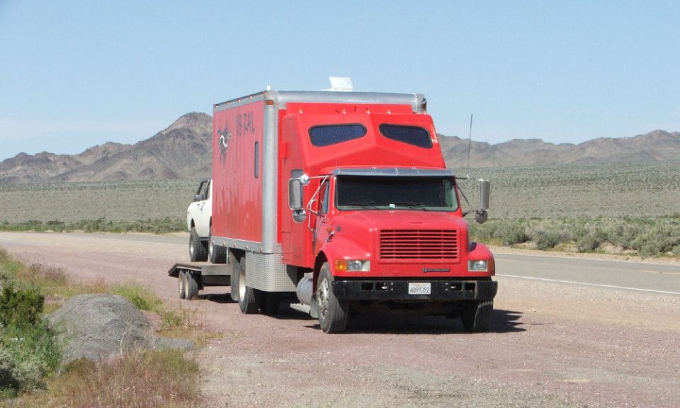 Red box truck towing a trailer loaded with a white pickup on a desert roadside.