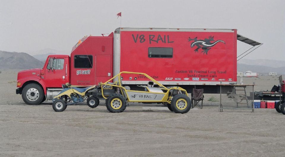 Red semi truck and 'V8 RAIL' trailer in desert with yellow sand rail buggy and blue ATV parked alongside