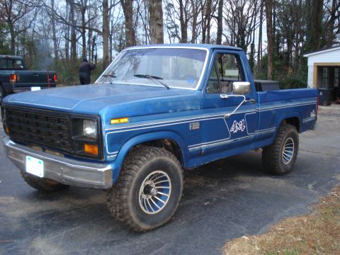 1985 Ford F-150 - A blue 1985 Ford F-150 4x4 pickup truck is parked on a driveway in front of a wooded area and a garage.
