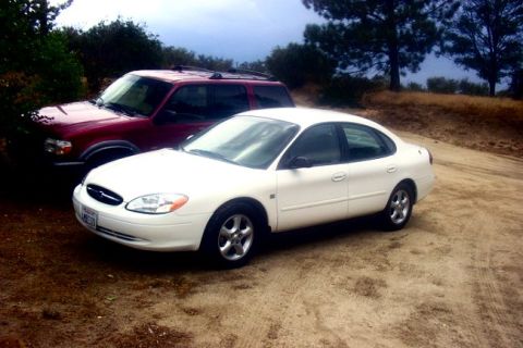 2000 Ford Taurus - A white 2000 Ford Taurus sedan is parked on a dirt road next to a red SUV.