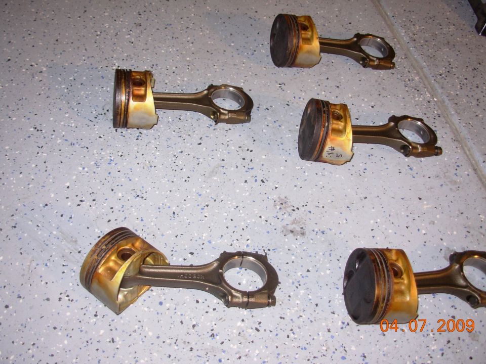 Several used pistons and connecting rods laid out on a speckled floor. Date stamp reads 04.07.2009