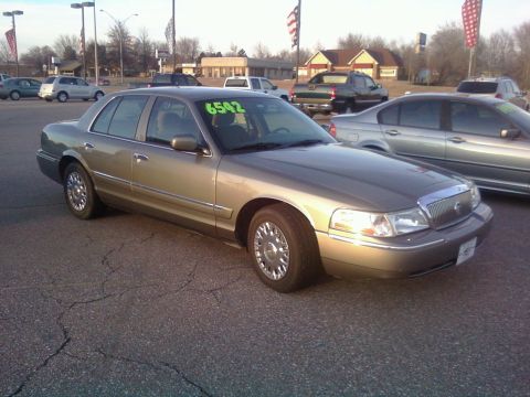 2000 Mercury Grand Marquis - A gold sedan with a chrome grille is parked in a lot, with other vehicles and buildings in the background.