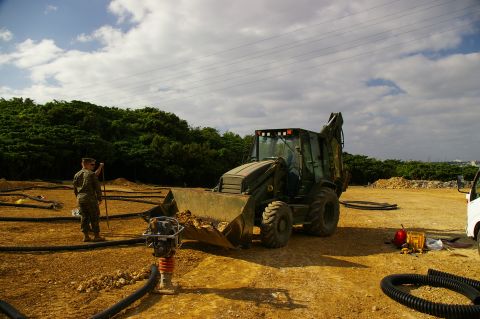 1986 Ford Bronco - A backhoe loader is positioned in a construction area, with a person observing the scene and various construction materials around.
