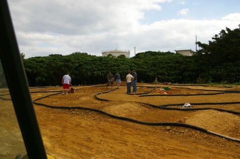 1986 Ford Bronco - A group of people stand on a dirt track designed for remote-controlled cars, with industrial tanks in the distance.