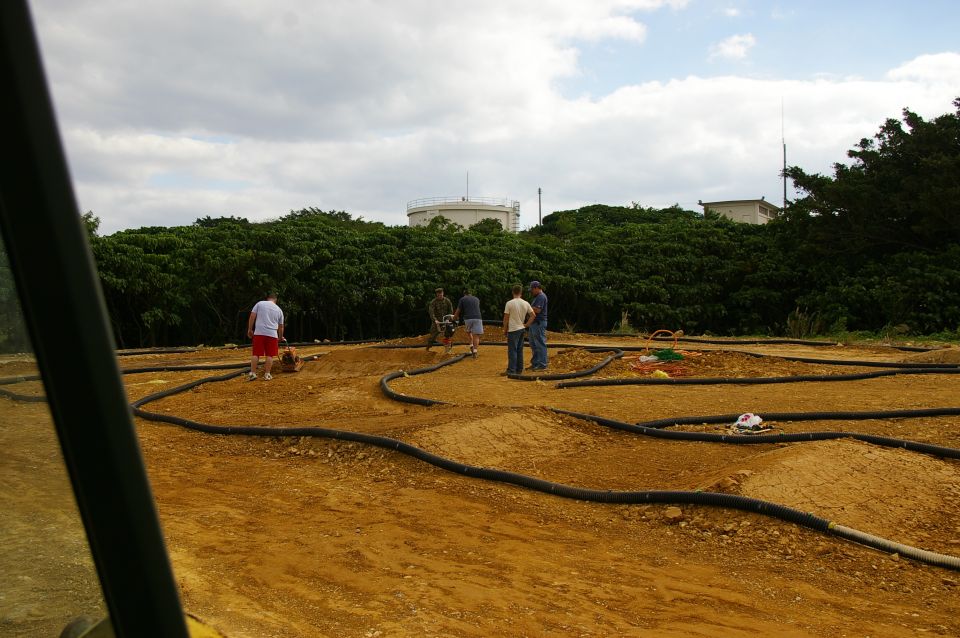 People gathered around a dirt track with black tubing outlining the course. Some industrial tanks visible in background