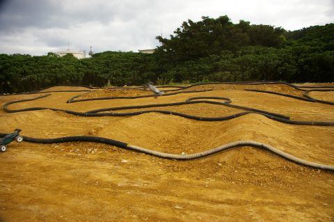 1986 Ford Bronco - Remote-controlled cars race on a dirt track with undulating terrain and black tubing borders.