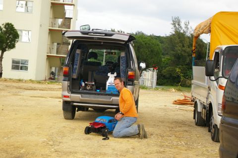 1986 Ford Bronco - A man kneels with remote control cars in front of a silver SUV with its rear hatch open, parked next to a white truck.