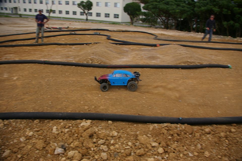 blue remote control car with red front driving on a dirt track with black tubing borders. Two people stand in background