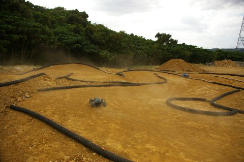 1986 Ford Bronco - RC cars race on a dusty dirt track with curves and jumps, surrounded by trees and construction debris.