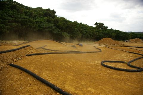 1986 Ford Bronco - A remote-controlled car races around a dusty dirt track bordered by black tubing, with a dense line of trees in the background.