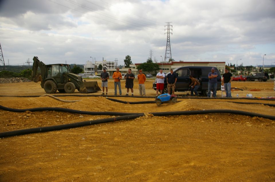 group of men watching remote control cars on a dirt track, with a backhoe and van in background