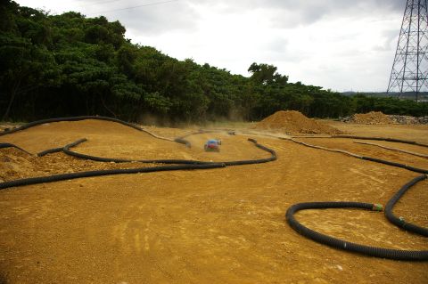 1986 Ford Bronco - RC cars race on a dusty dirt track with tubing borders and trees in the background.
