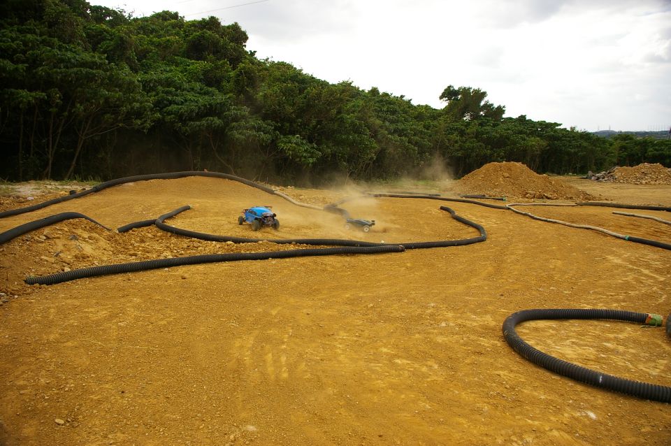Two remote control cars race on a dirt track with black tubing for boundaries. Trees are in the background
