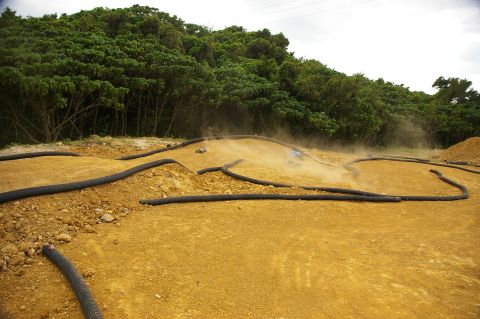 1986 Ford Bronco - Remote control cars race on a dusty track bordered by black tubing, with a dense treeline behind them.
