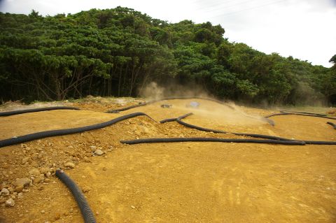 1986 Ford Bronco - A remote control car races on a dusty track, bordered by black tubing, with a dense forest in the background.