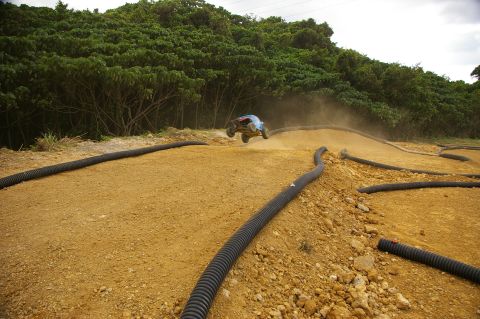 1986 Ford Bronco - A remote controlled car leaps over a dirt track obstacle, kicking up dust.