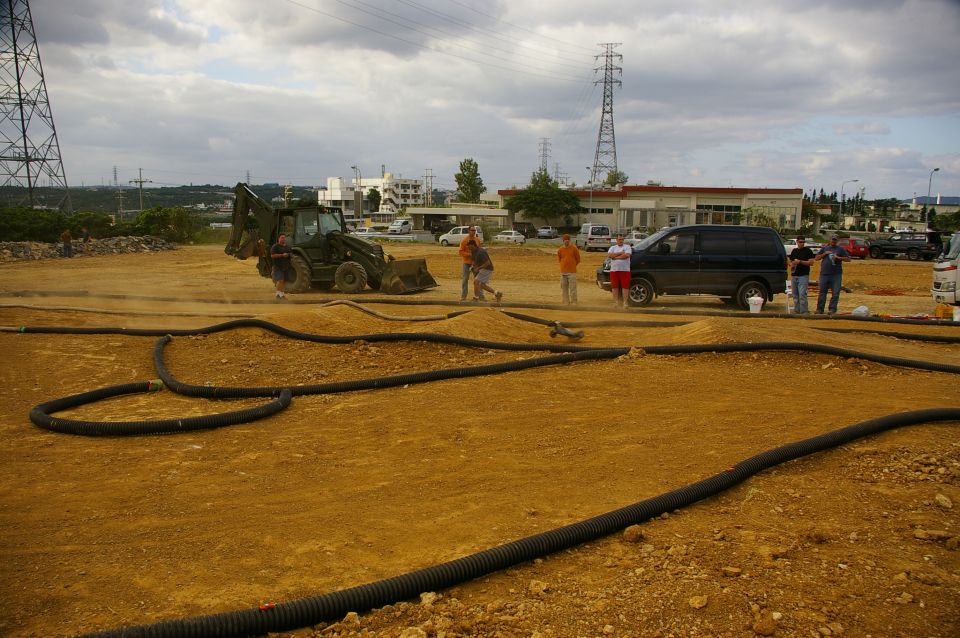 RC car track with black tubing, dirt mounds, a backhoe, a van, and people standing around