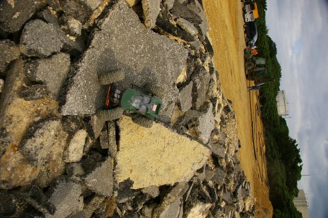 1986 Ford Bronco - RC rock crawler toy on debris, with construction vehicles and tanks in background.