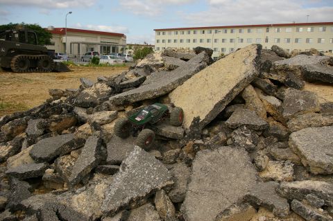 1986 Ford Bronco - A remote control rock crawler navigates a mound of broken asphalt, with buildings in the background.