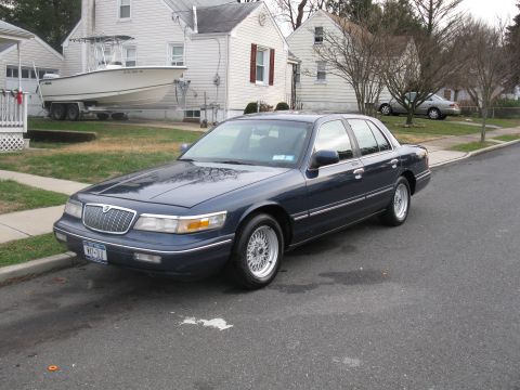 1995 Mercury Grand Marquis - A dark blue Mercury Grand Marquis is parked on a residential street with a boat on a trailer in the driveway.