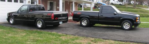 1990 Chevrolet Silverado 1500 - Two black single-cab, short-bed Chevrolet 1500 pickups parked nose-to-tail on a residential driveway. One shows its tailgate with a '454 SS…