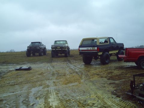 1990 Ford Bronco - A lifted 1990 Ford Bronco is parked in a muddy field alongside two other trucks and a red truck bed.