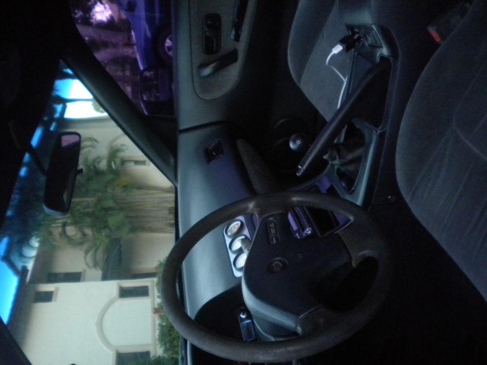 Car interior with steering wheel, dashboard gauges, and center console. A phone is charging