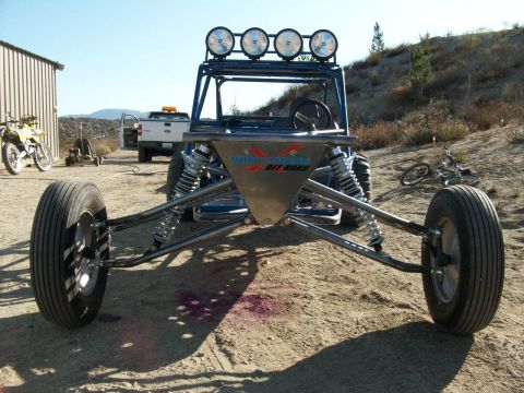 1996 Ford Bronco - An off-road buggy with a roll cage and four spotlights on top sits in a sandy area next to a building and a motorcycle.