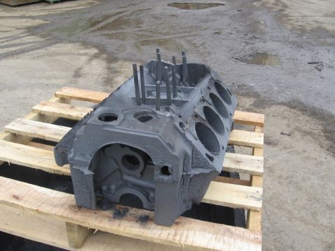 1972 Ford Ranchero - Rough as-cast V8 engine block sitting on a wood pallet outdoors on wet pavement, photographed from the front-top corner with unmachined cyl…