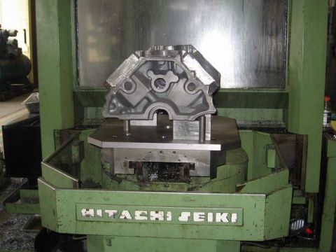 1972 Ford Ranchero - A V-shaped iron engine block casting sits front-face-up on a fixture plate inside a green Hitachi Seiki machining center, with the cam bore…