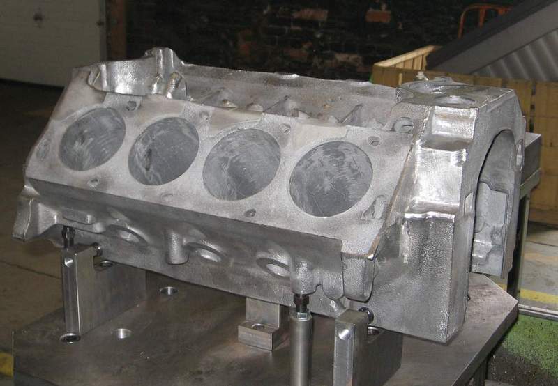 Rough-cast V8 block fixtured on a machining plate, four unmachined bores facing the camera