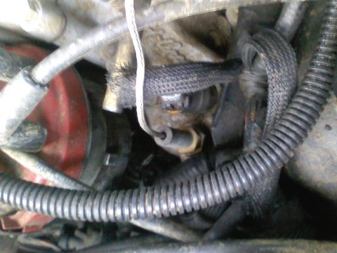 1990 Ford Bronco - Tight engine-bay close-up of a grimy wiring harness in braided sleeving and split-loom conduit routed past a black electrical connector and…