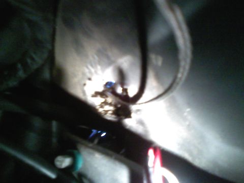 1990 Ford Bronco - Dark, blurry flash close-up under a vehicle showing a rusty hole or corroded penetration in a sheet-metal panel with a wire or cable routed…