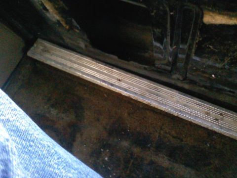 1990 Ford Bronco - Close-up of a truck's door sill with the ribbed aluminum scuff plate in place, looking down at a rusty, dirt-covered cab floor with the car…