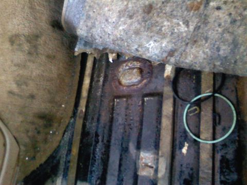 1990 Ford Bronco - Tight close-up of a dirty, ribbed sheet-metal floor pan under a vehicle, showing a rusted circular boss or plug in the stamping with surfac…