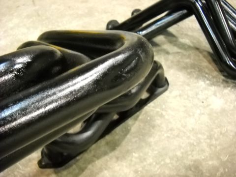 1978 Ford Bronco - Close-up of a pair of black-coated tubular exhaust headers laid out on a bare concrete floor, showing the flange and the tight bends where…