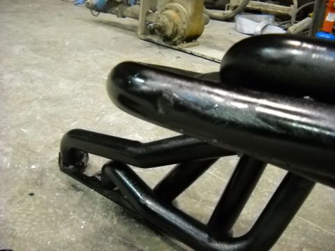 1978 Ford Bronco - Freshly painted gloss-black tubular bumper or grille guard resting on a shop floor, shot low and close along its length. The fabricated rou…