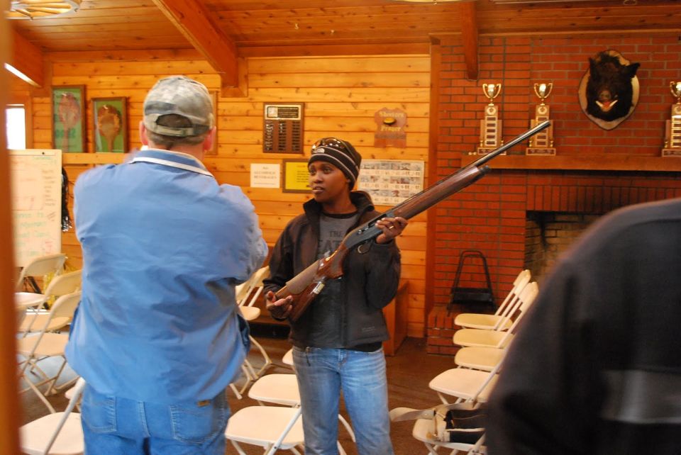 Two people in a room with wood paneling and chairs, one holding a shotgun