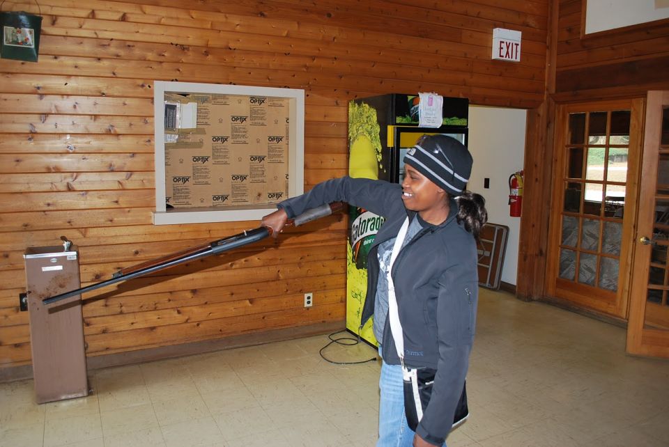 Woman in a beanie and jacket holds a rifle, aiming it towards a water fountain. A vending machine is behind her