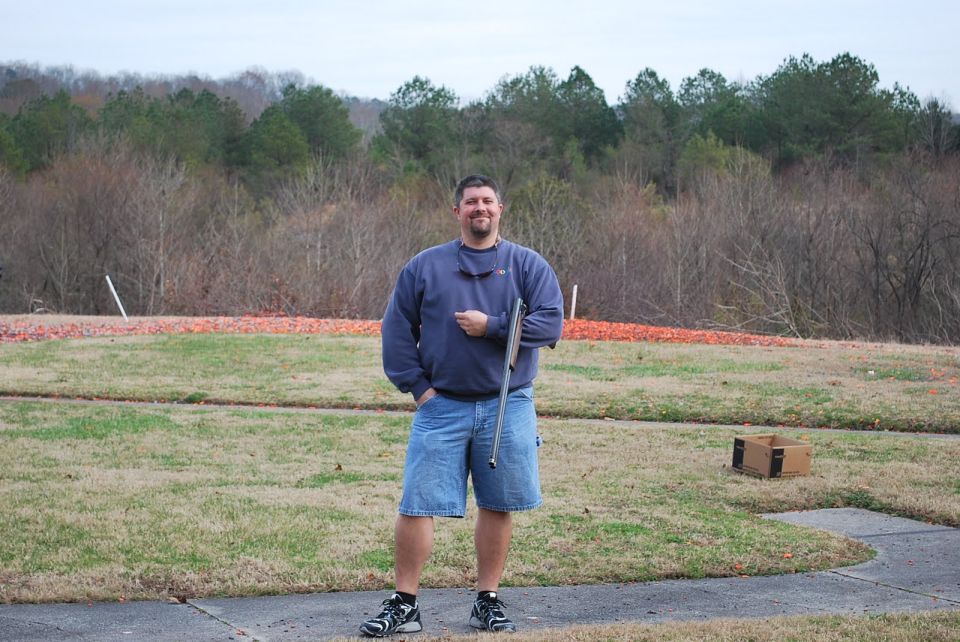 Man in a blue sweatshirt and denim shorts stands in a grassy field, holding a rifle. Trees and a cloudy sky are in the background