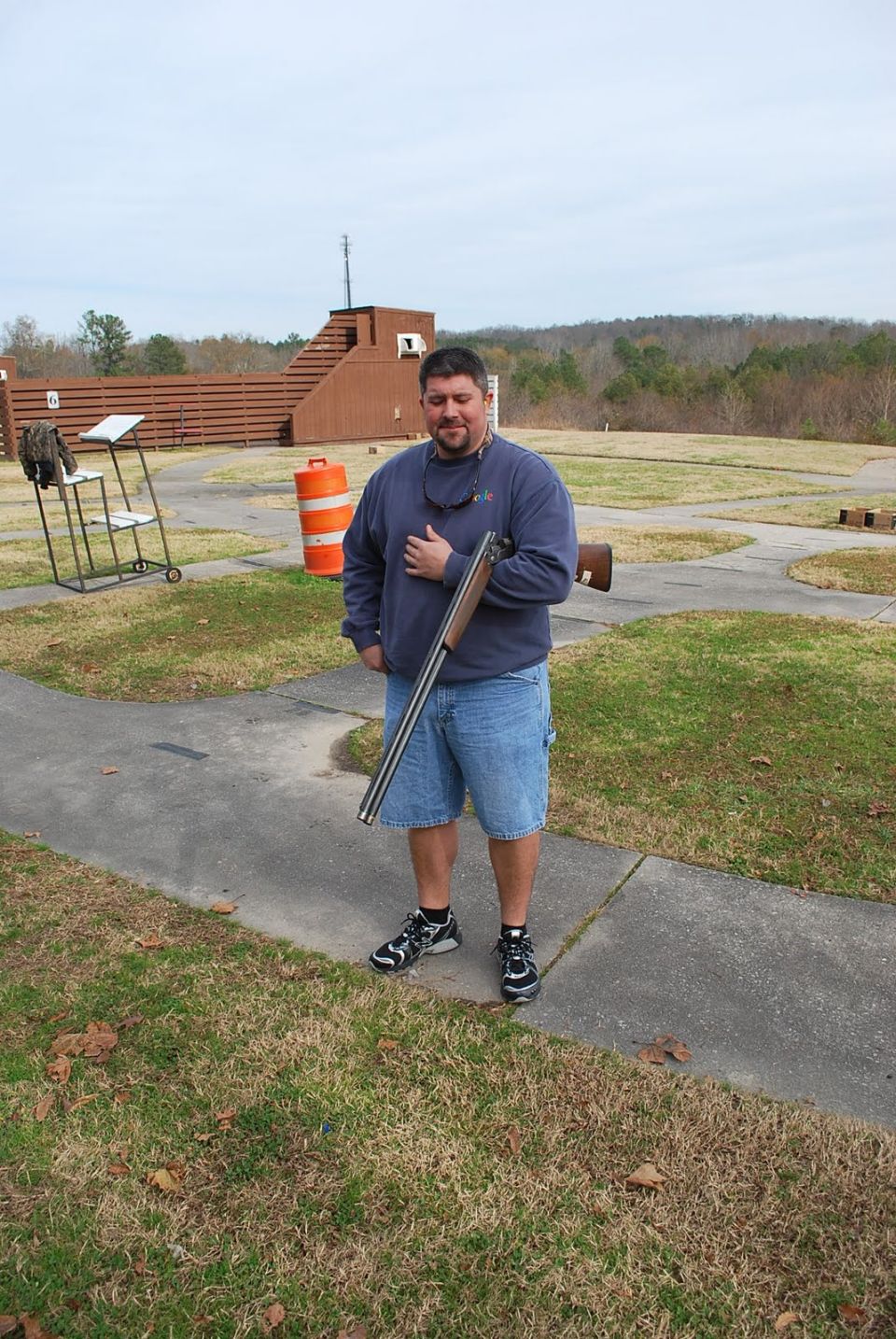 Man in blue sweatshirt and denim shorts holding a shotgun at a shooting range